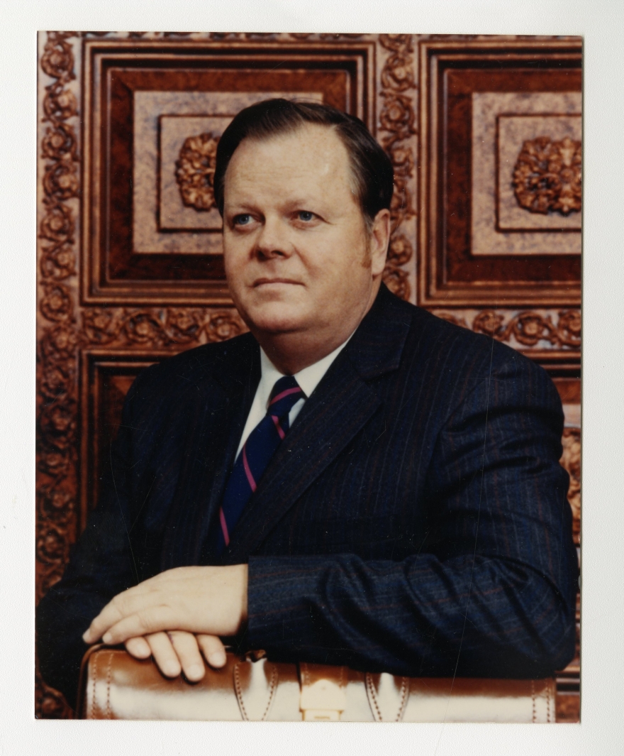 Ron Campbell in a formal pose wearing a navy suit with an ornate wooden panel background. background and a